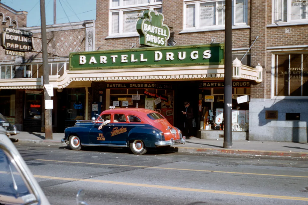 A cream and maroon taxi on Greenwood Avenue in Seattle in the 1950s, parked in front of a Bartell Drugs storefront.