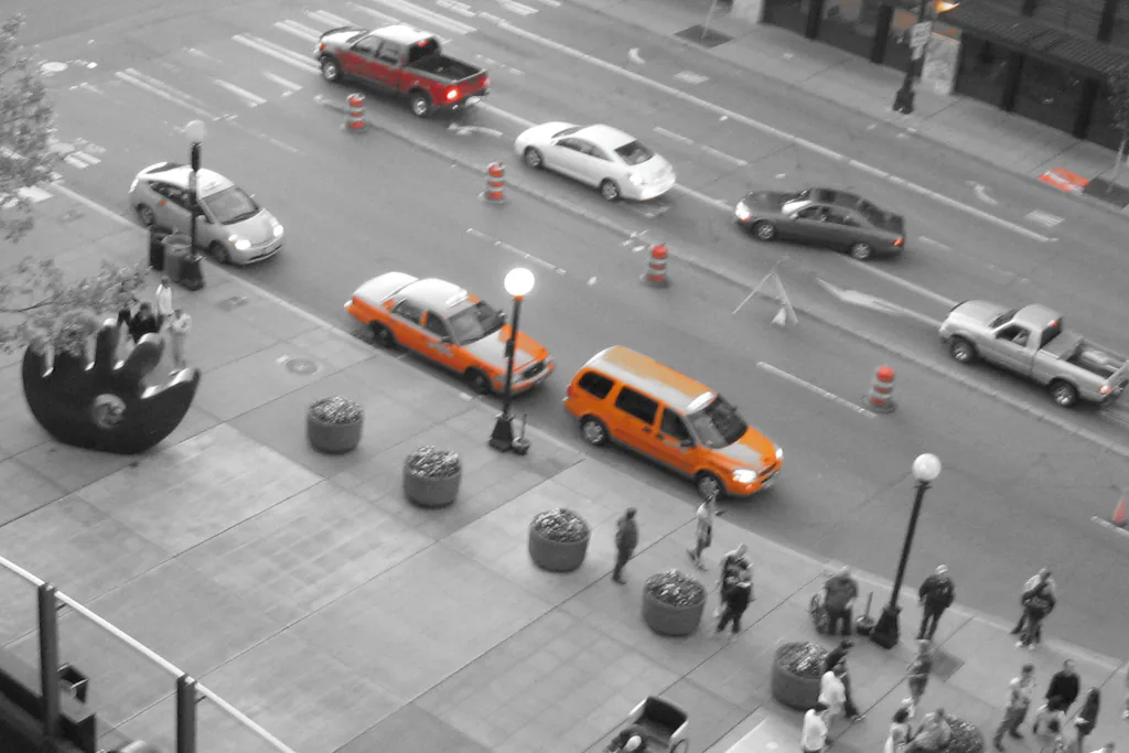 Looking down on a downtown Seattle street: an orange saloon taxi with a roof sign and an orange taxi minivan wait one behind the other at the kerb while pedestrians pass on the pavement. The photograph is desaturated apart from the cabs, a red pickup crossing the road and the orange traffic cones.