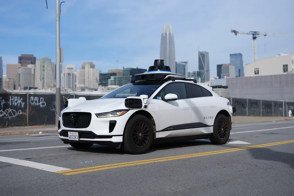 A white Jaguar I-Pace with a roof-mounted sensor pod stands at a San Francisco kerb, the downtown skyline rising behind it.